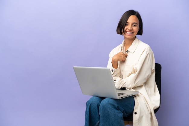 Mulher jovem de raça mista sentada em uma cadeira com laptop isolado em um fundo roxo com expressão facial surpresa