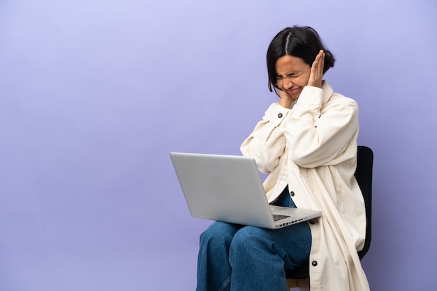 Mulher jovem de raça mista sentada em uma cadeira com laptop isolado em um fundo roxo frustrada e cobrindo as orelhas