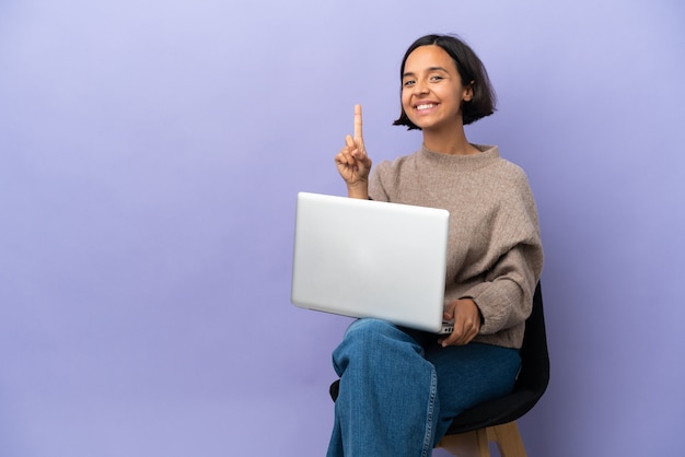 Mulher jovem de raça mista sentada em uma cadeira com laptop isolado em um fundo roxo, mostrando e levantando um dedo