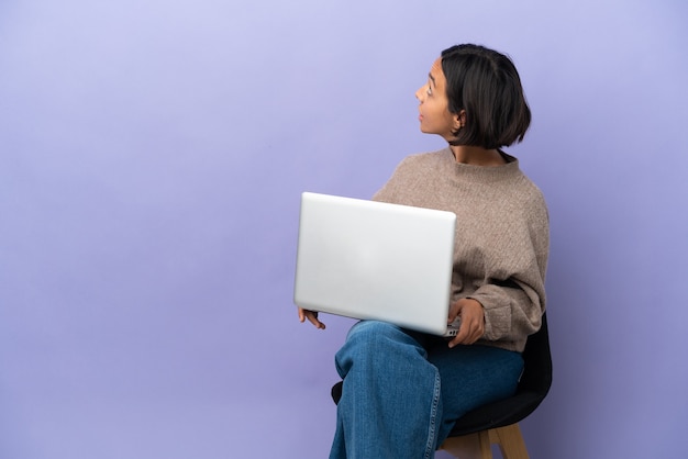 Mulher jovem de raça mista sentada em uma cadeira com laptop isolado em um fundo roxo na posição traseira e olhando para trás