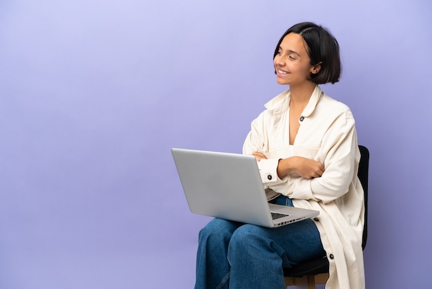 Mulher jovem de raça mista sentada em uma cadeira com laptop isolado em um fundo roxo, olhando para o lado e sorrindo