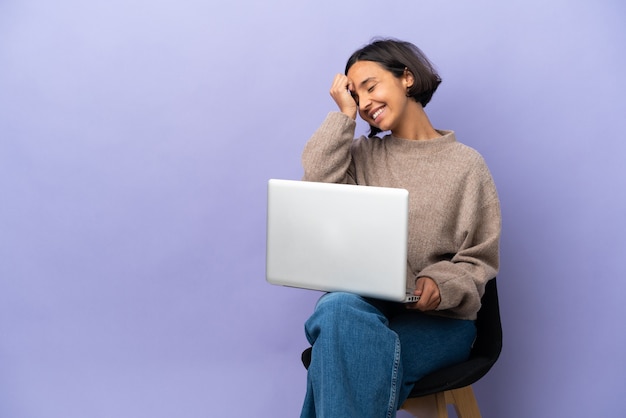 Mulher jovem de raça mista sentada em uma cadeira com laptop isolado em um fundo roxo sorrindo muito