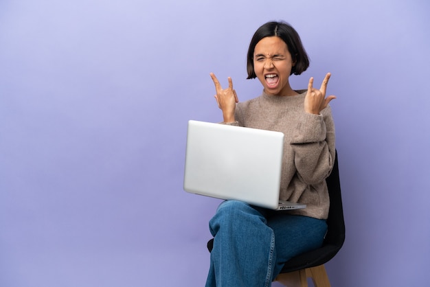 Foto mulher jovem de raça mista sentada em uma cadeira com laptop isolado no fundo roxo fazendo gesto de chifre