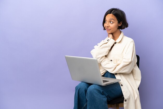 Mulher jovem de raça mista sentada em uma cadeira com laptop isolado no fundo roxo pensando uma ideia