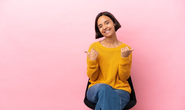 Mulher jovem de raça mista sentada em uma cadeira isolada em um fundo rosa com gesto de polegar para cima e sorrindo