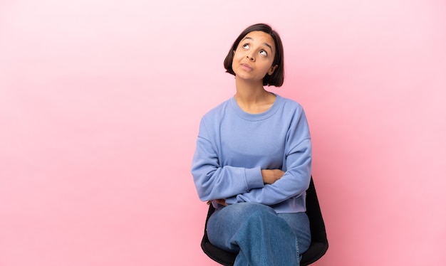 Mulher jovem de raça mista sentada em uma cadeira isolada em um fundo rosa e olhando para cima