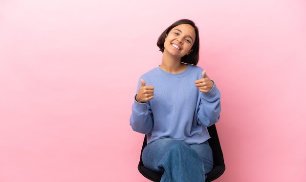 Mulher jovem de raça mista sentada em uma cadeira isolada em um fundo rosa fazendo um gesto de polegar para cima