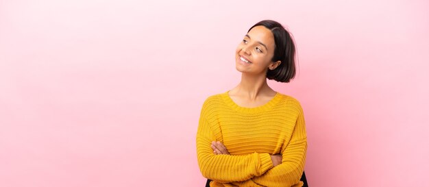 Mulher jovem de raça mista sentada em uma cadeira isolada em um fundo rosa feliz e sorridente