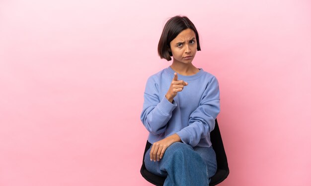 Mulher jovem de raça mista sentada em uma cadeira isolada em um fundo rosa frustrada e apontando para a frente