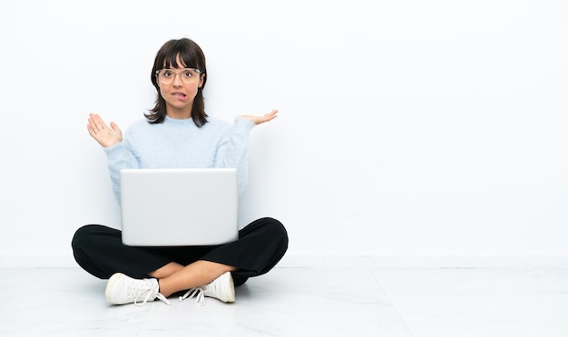 Mulher jovem de raça mista sentada no chão com laptop isolado no fundo branco com dúvidas ao levantar as mãos