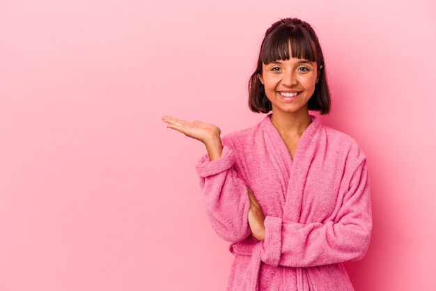 Mulher jovem de raça mista, vestindo um roupão de banho isolado no fundo rosa, mostrando um espaço de cópia na palma da mão e segurando a outra mão na cintura.