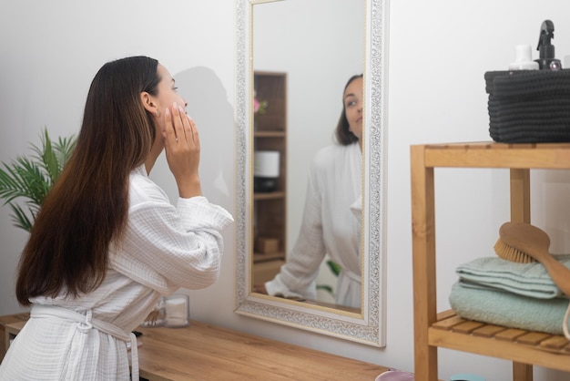 Mulher jovem de roupão de banho olhando para o espelho na casa de banho senhora bonita tocando pele facial saudável