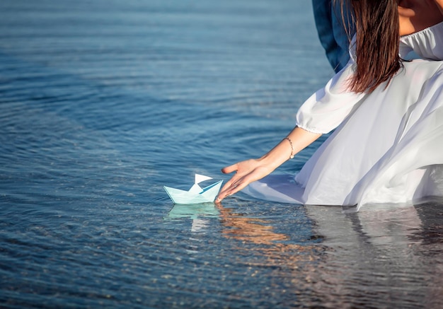 Mulher jovem de vestido branco segura um barco de papel azul claro na mão em fundo do mar