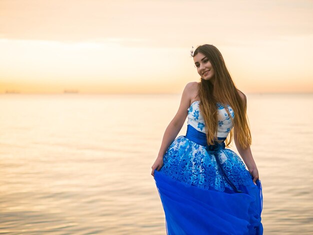 Mulher jovem de vestido de pé contra o mar durante o pôr-do-sol