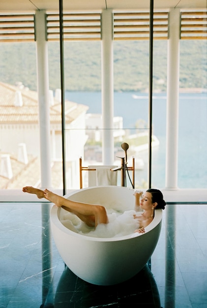 Mulher jovem deitada em uma banheira redonda com espuma segurando as bordas em um hotel com colunas e vista para o mar