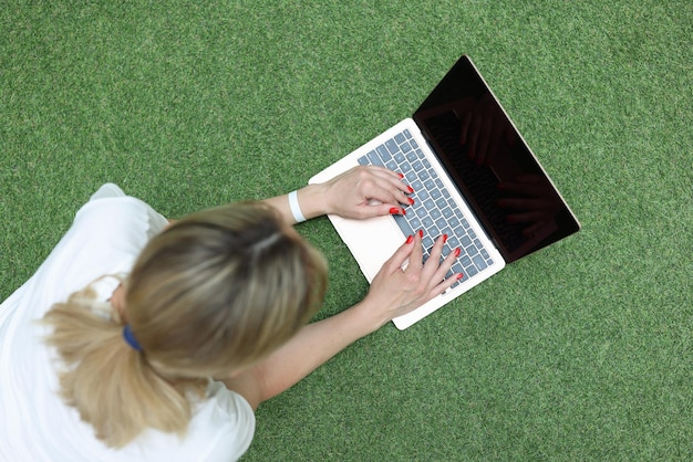 Mulher jovem deitada na grama verde e digitando na vista superior do teclado do laptop