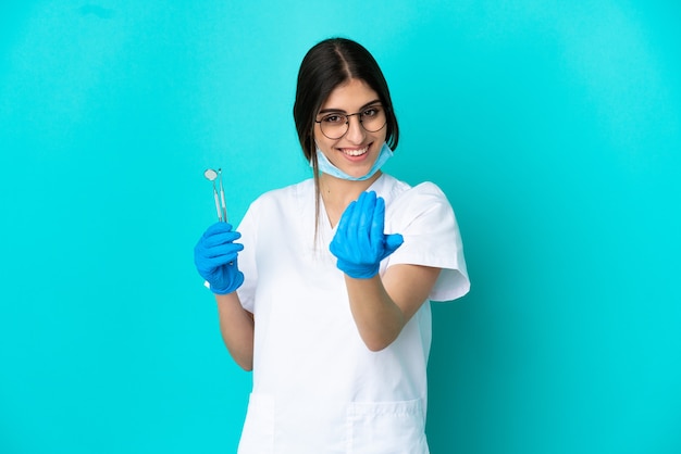 Mulher jovem dentista caucasiana segurando ferramentas isoladas sobre fundo azul, convidando para vir com a mão. Feliz que você veio