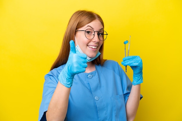 Mulher jovem dentista ruiva isolada em fundo amarelo com polegares para cima porque algo bom aconteceu