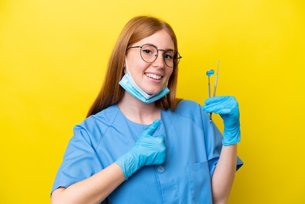 Mulher jovem dentista ruiva isolada em fundo amarelo dando um polegar para cima gesto