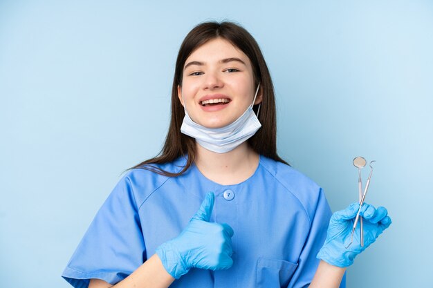 Mulher jovem dentista segurando ferramentas sobre parede azul isolada, dando um polegar para cima gesto