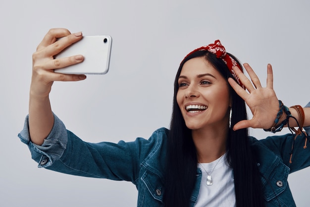 Mulher jovem descolada em bandana fazendo selfie e sorrindo em pé contra uma parede cinza