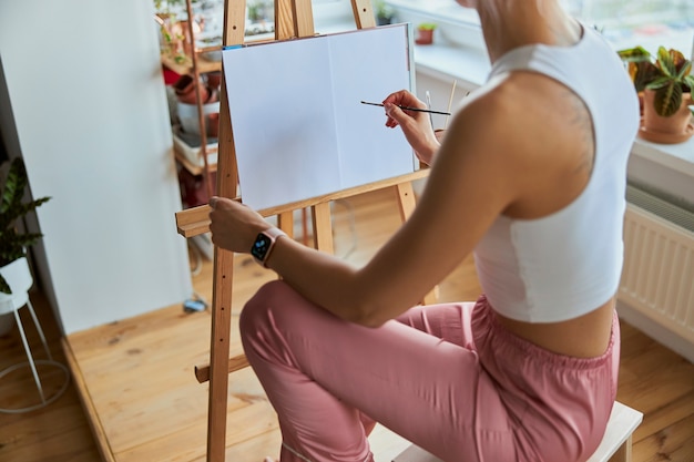 Mulher jovem desenha imagens na tela com o pincel no cavalete de madeira na varanda