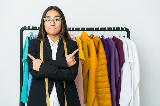 Mulher jovem designer asiática isolada na parede branca aponta para o lado, está tentando escolher entre duas opções.