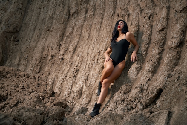 Mulher jovem deslumbrante em um bodysuit preto com pernas longas esguias, em pé entre a pedreira de areia. senhora sexy com maquiagem brilhante e longos cabelos escuros, posando na câmera ao ar livre.