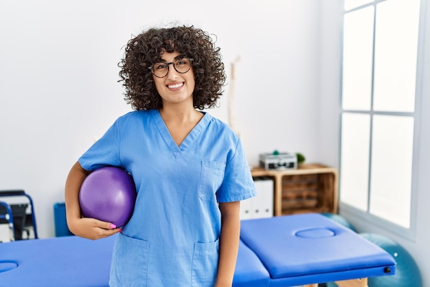 Mulher jovem do oriente médio vestindo uniforme de fisioterapeuta segurando bola em forma na clínica