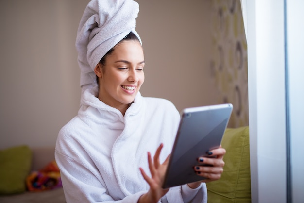 Mulher jovem e aconchegante alegre está usando tablet enquanto usava roupão e toalha de papel sobre a cabeça dela.