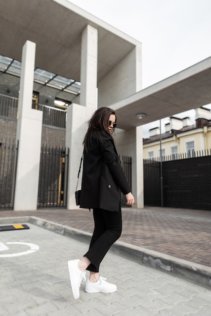 Mulher jovem e adorável elegante no blazer da juventude vintage em óculos de sol da moda com bolsa de couro se passando perto de um edifício moderno na cidade. Hipster de garota muito na moda legal em jaqueta preta da moda ao ar livre