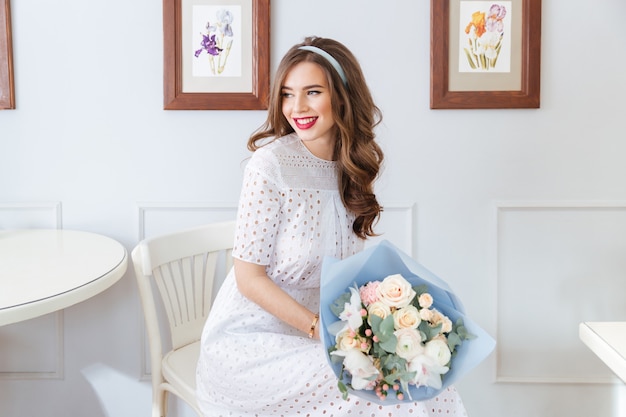Mulher jovem e adorável feliz em um vestido branco com buquê de flores sentada em um café
