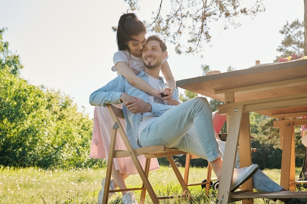 Mulher jovem e afetuosa feliz com um copo de vinho tinto abraçando seu namorado sorridente, sentado na cadeira ao lado da mesa servida ao ar livre