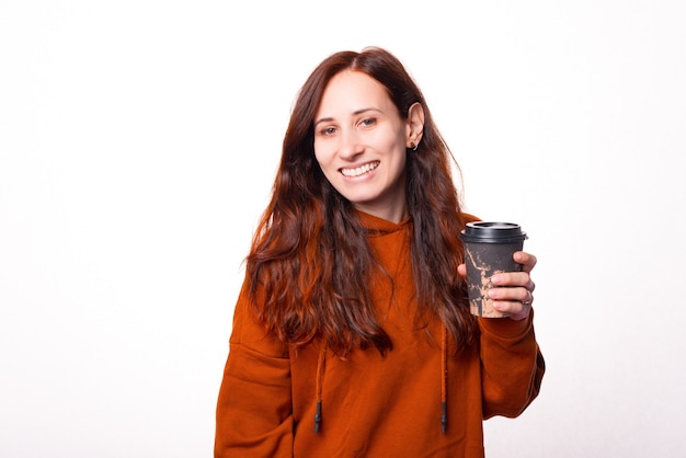 Mulher jovem e alegre está sorrindo para a câmera e segurando um copo com uma bebida quente