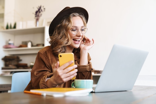 Mulher jovem e animada com chapéu, trabalhando com um laptop e um telefone celular, enquanto está sentado à mesa