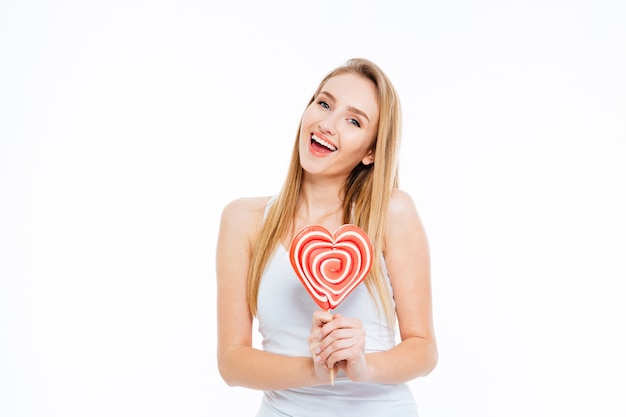 Mulher jovem e animada feliz em pé e segurando um pirulito em forma de coração sobre fundo branco