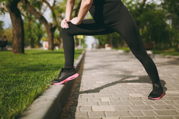 Mulher jovem e atlética, recortada, de uniforme preto, fazendo exercícios de alongamento, aquecimento antes de correr ou treinar, em pé no parque da cidade ao ar livre
