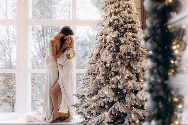 mulher jovem e atraente abraçando o namorado sem camisa perto da árvore de natal em casa