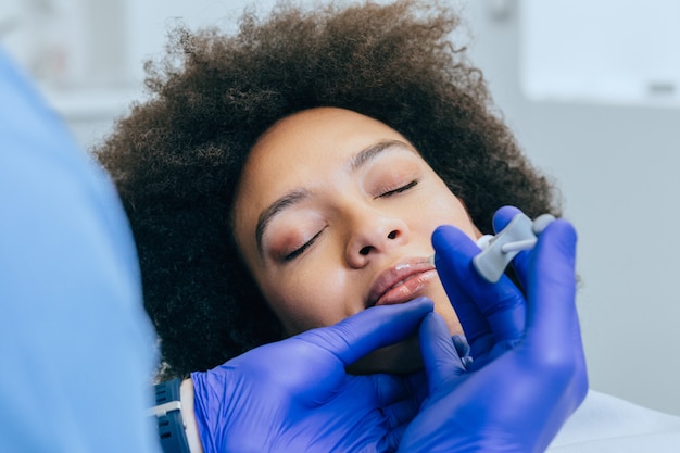 Mulher jovem e atraente africana está recebendo injeções faciais rejuvenescedoras. Ela está calmamente sentada na clínica.