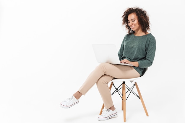 Foto mulher jovem e atraente africana sentada em uma cadeira isolada sobre uma parede branca, usando um laptop