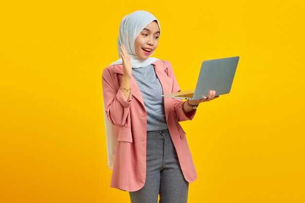 Mulher jovem e atraente asiática sorrindo segurando um laptop e cumprimentando sobre fundo amarelo