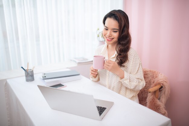 Mulher jovem e atraente asiática usando laptop enquanto bebe chá / café.