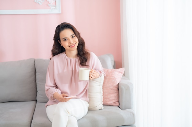 Mulher jovem e atraente asiática usando telefone e bebendo café quando está sentado no sofá em casa.