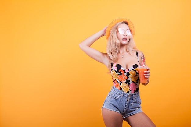 Mulher jovem e atraente bebendo suco de laranja com óculos de sol no estúdio