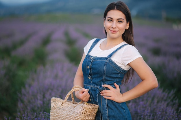 Mulher jovem e atraente caminhando no campo de lavanda de verão Modelo vestida com vestido de verão jeans com chapéu de palha e bolsa