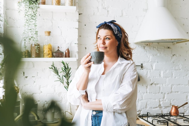 Mulher jovem e atraente com corpo de tamanho positivo em jeans azul e camisa branca bebe café da manhã