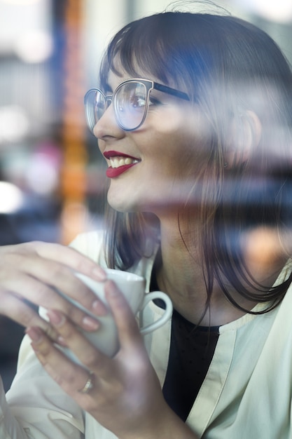 Foto mulher jovem e atraente com lábios vermelhos usa óculos, olhando pela janela e bebe café no restaurante