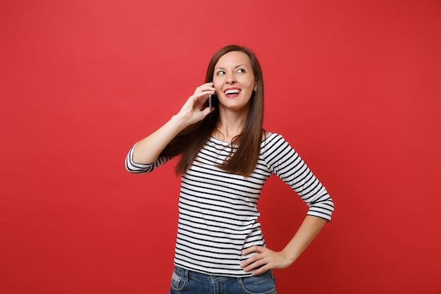 Mulher jovem e atraente com roupas listradas, falando no celular, conduzindo uma conversa agradável isolada sobre fundo vermelho brilhante. Emoções sinceras de pessoas, conceito de estilo de vida. Simule o espaço da cópia.