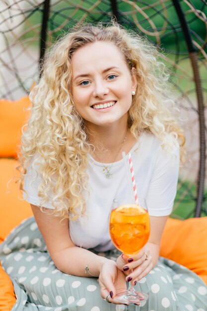 Mulher jovem e atraente com sorriso encantador tem cabelos ondulados vestidos casualmente mantém o coquetel de verão fresco recria no jardim durante o fim de semana Conceito de descanso e estilo de vida de pessoas Tiro vertical