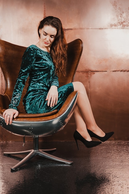 Mulher jovem e atraente com vestido de veludo verde sentada em uma cadeira de couro marrom